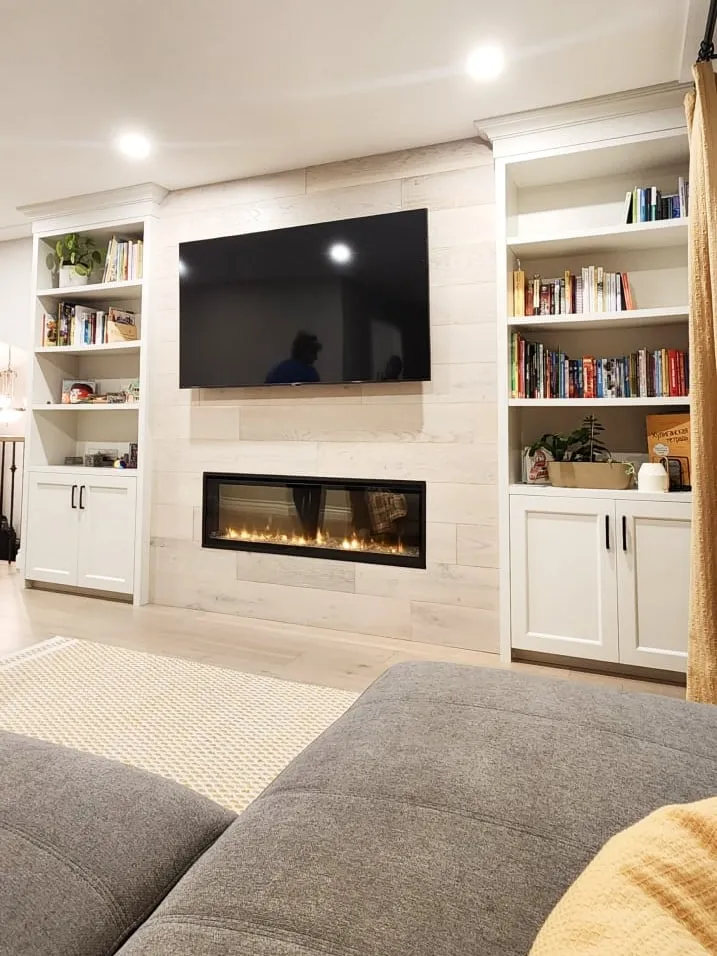 A finished living room seen from a grey sofa, facing a lit fireplace wall